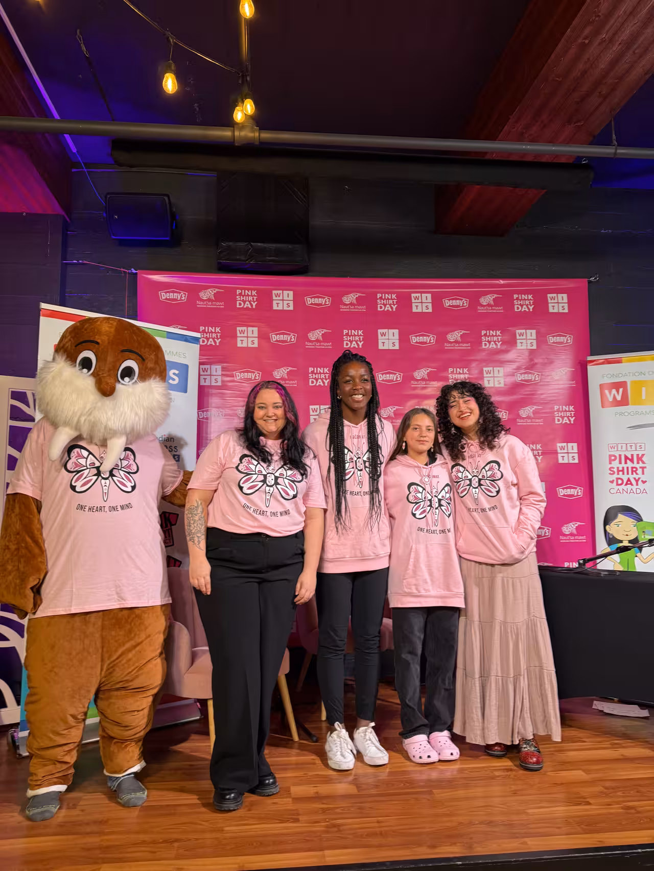 Four people and the WITSup walrus mascot, all wearing pink shirts, pose together in front of a Pink Shirt Day backdrop