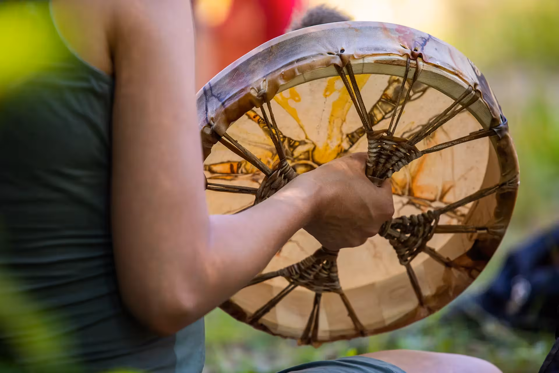 A person holding a traditional rawhide hand drum, showing the intricate lacing on the back and a yellow painted design on the drumhead.