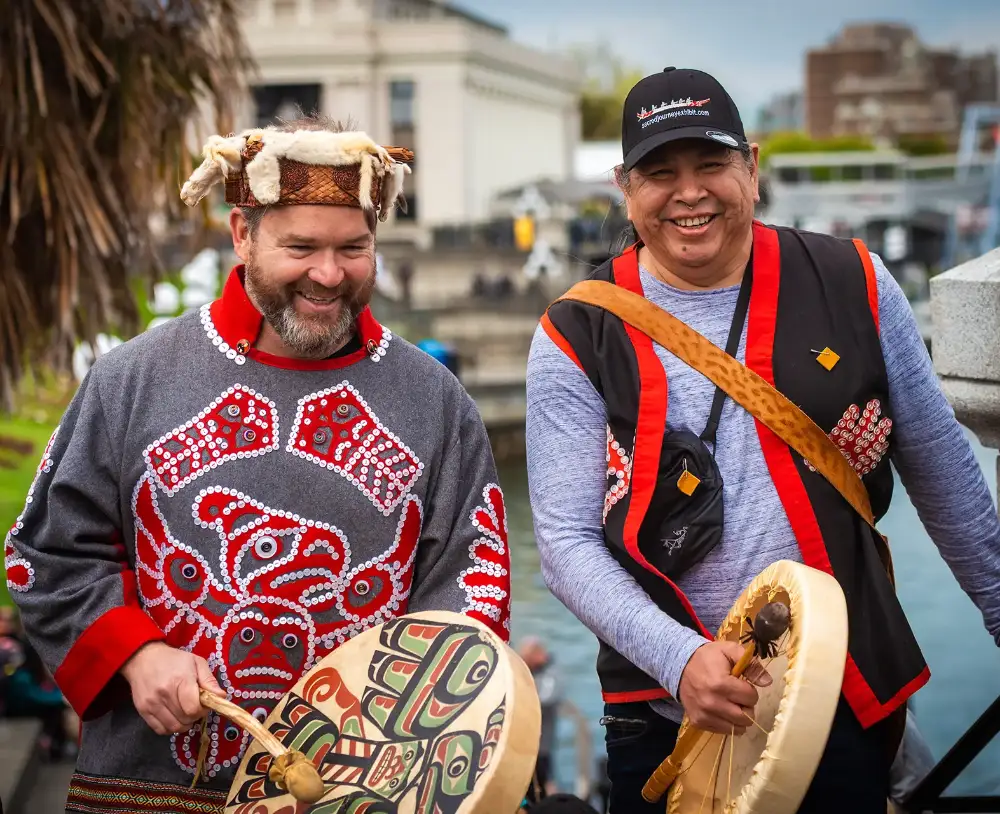 Two men in traditional regalia holding hand drums and smiling